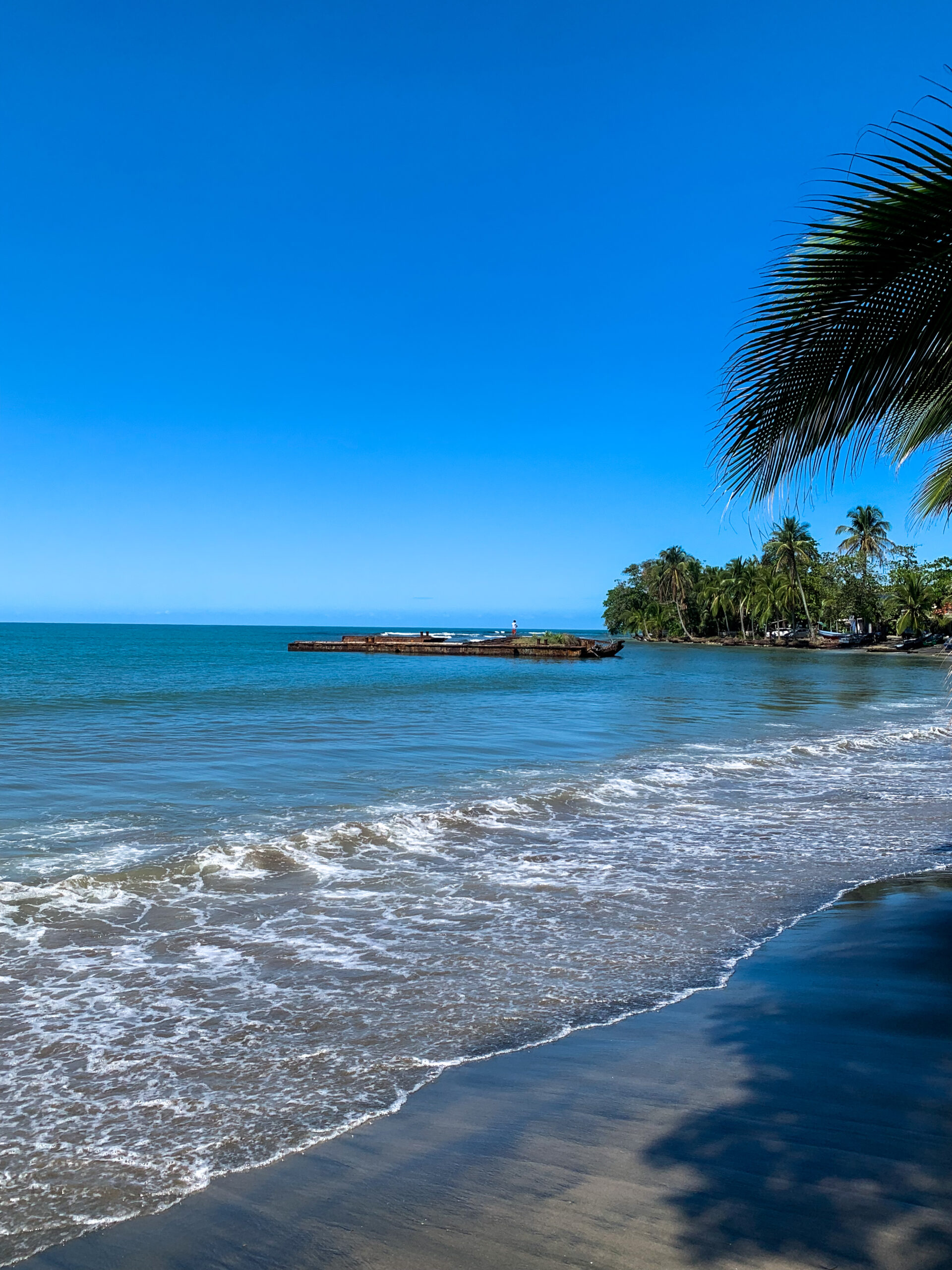 Image of the ocean and sand at Playa Negra in Puerto Viejo Costa Rica. Old shipwreck in the water and trees.